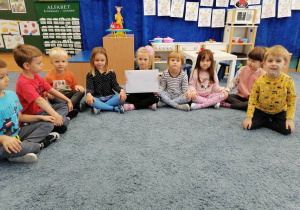 Grupa dzieci siedzi na dywanie; dziewczynka trzyma kopertę.